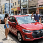 Fotografia de rua no Brasil mostrando um homem negro sorridente segurando as chaves de um carro hatchback popular vermelho metálico recém-adquirido, ao lado de uma mulher negra na calçada.